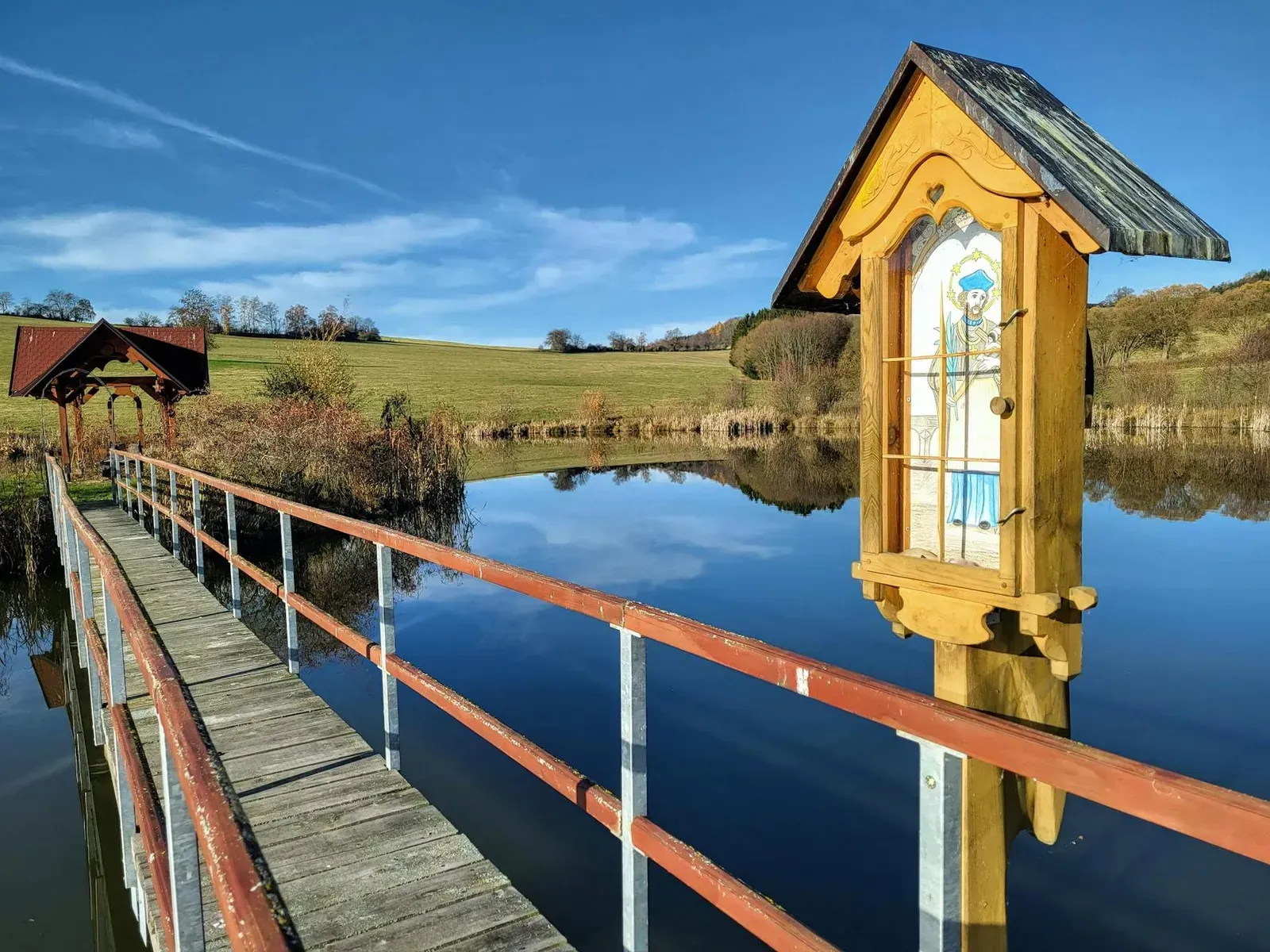 Tranquil scene of a wooden shrine by a pond in Jihočeský kraj, Czech Republic, reflecting blue skies.